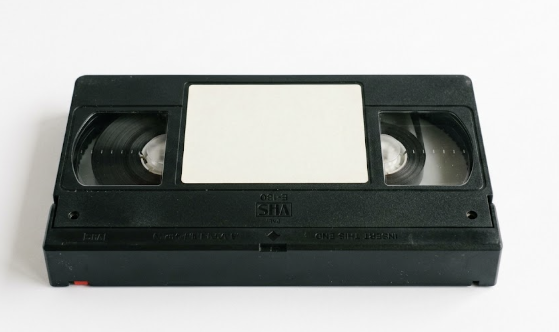 Casete VHS apoyado sobre fondo claro, con la etiqueta todavia en blanco y las dos bobinas de cinta visibles tras las ventanas de plastico.